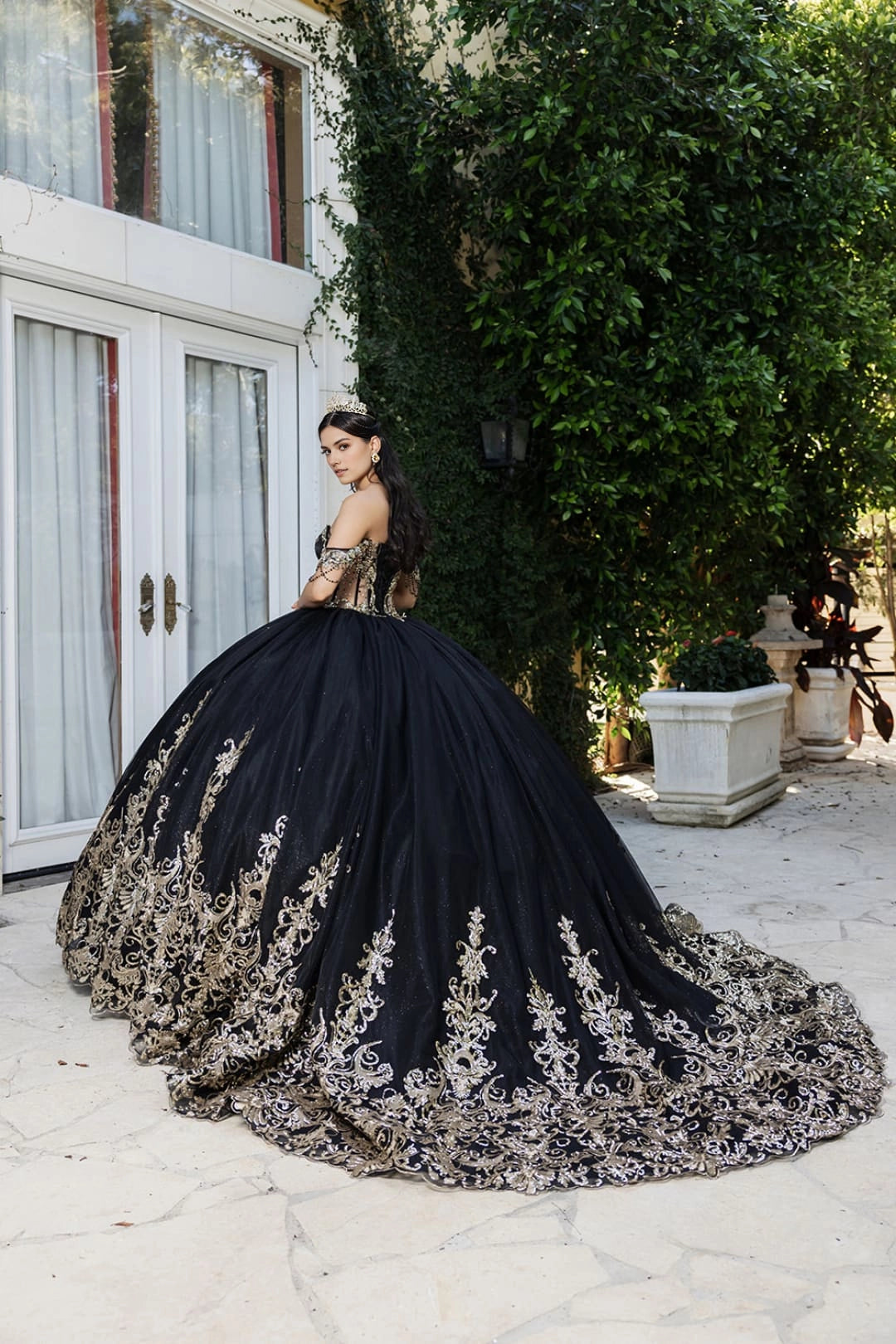 Rear three-quarter view of Vedress black quinceañera ball gown with gold-toned scrollwork along the full skirt and long scalloped train.